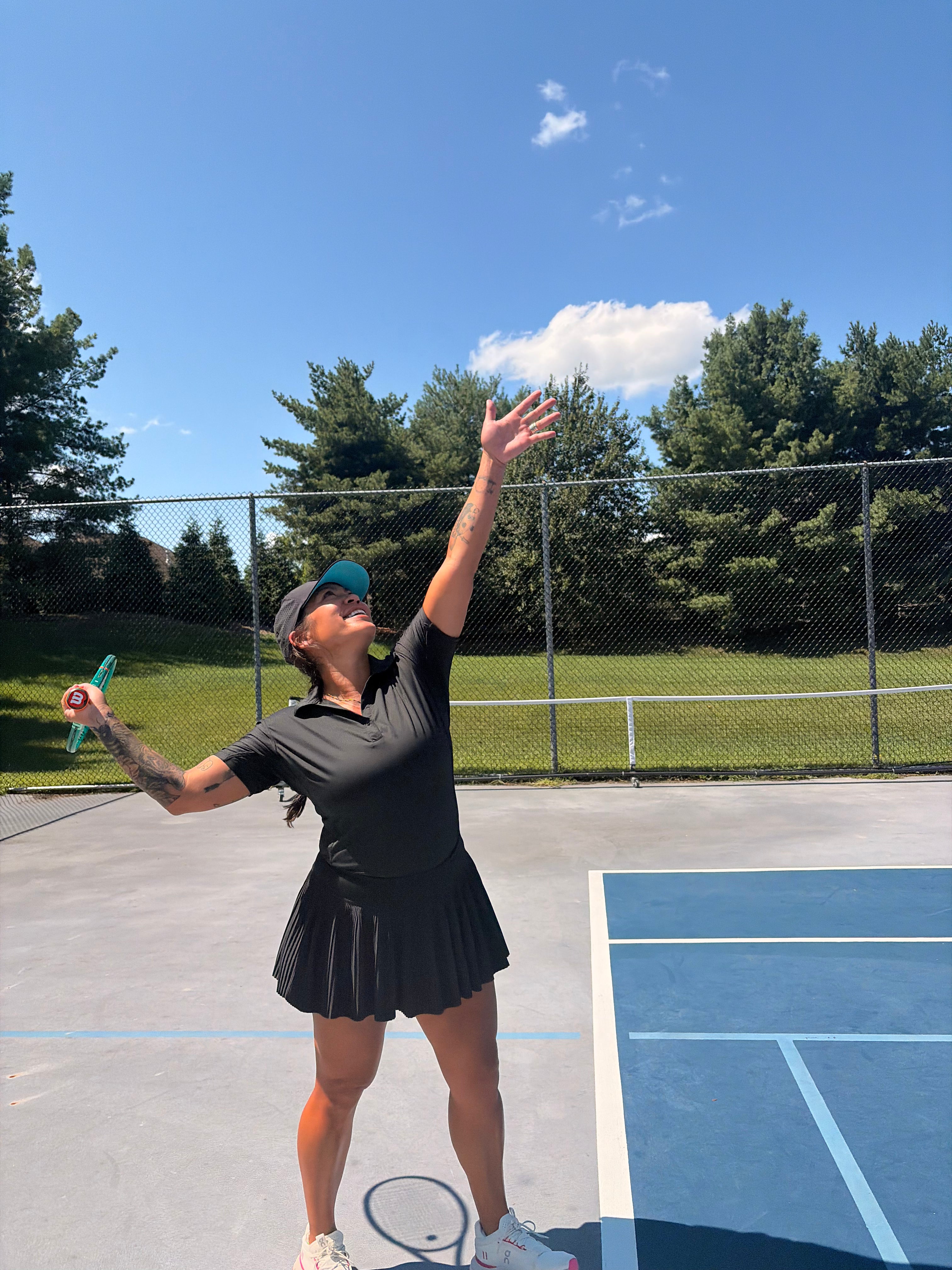 A woman in a black tennis outfit and visor serves on an outdoor tennis court under a blue sky with trees in the background.