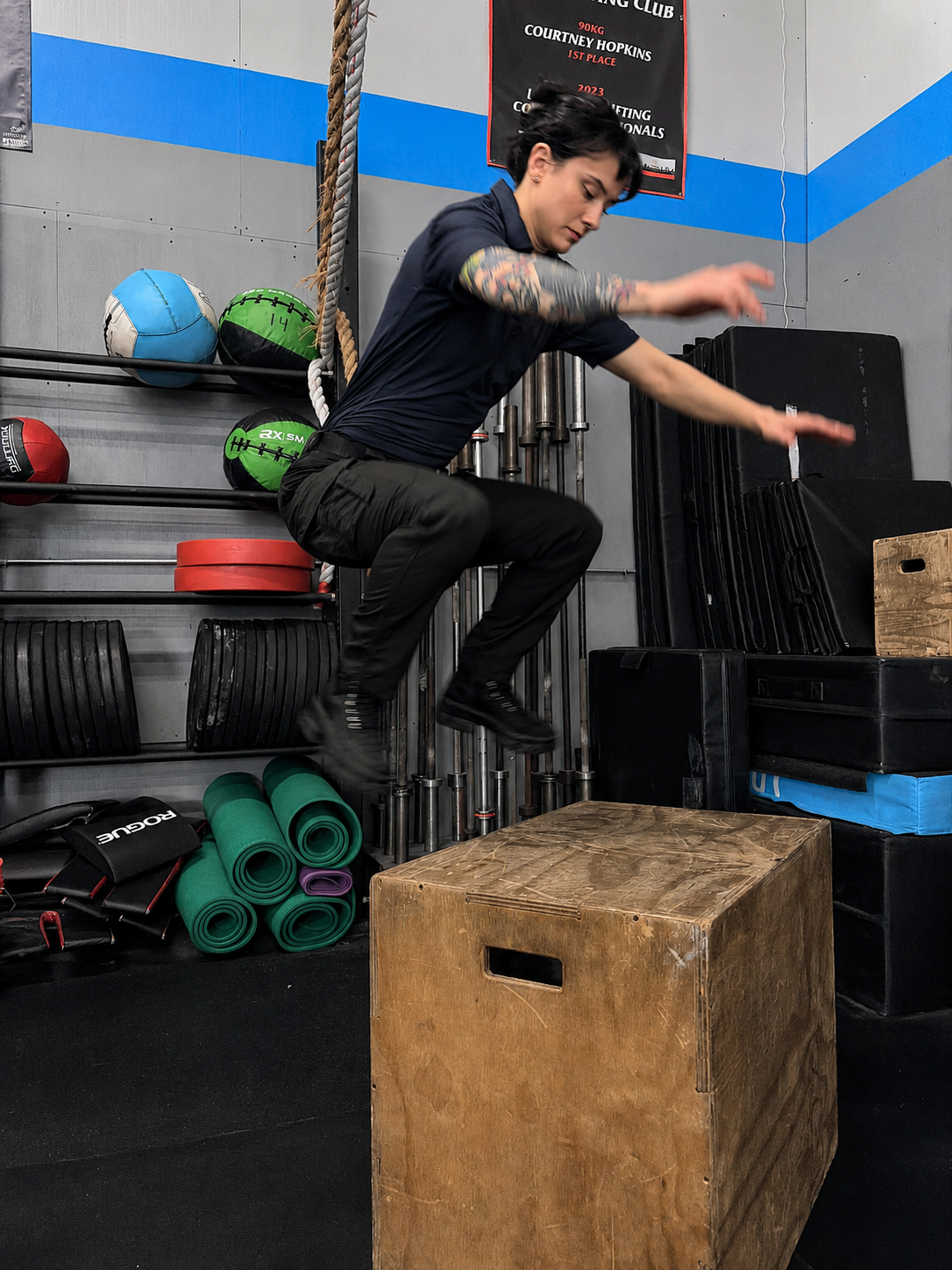A man with tattoos jumps onto a wooden plyo box in a gym setting, wearing a navy blue t-shirt and black cargo pants. The background contains exercise equipment, weight plates, and mats.