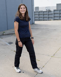 Smiling woman wearing black MGT Gear workwear pants outdoors, full-length angled front view
