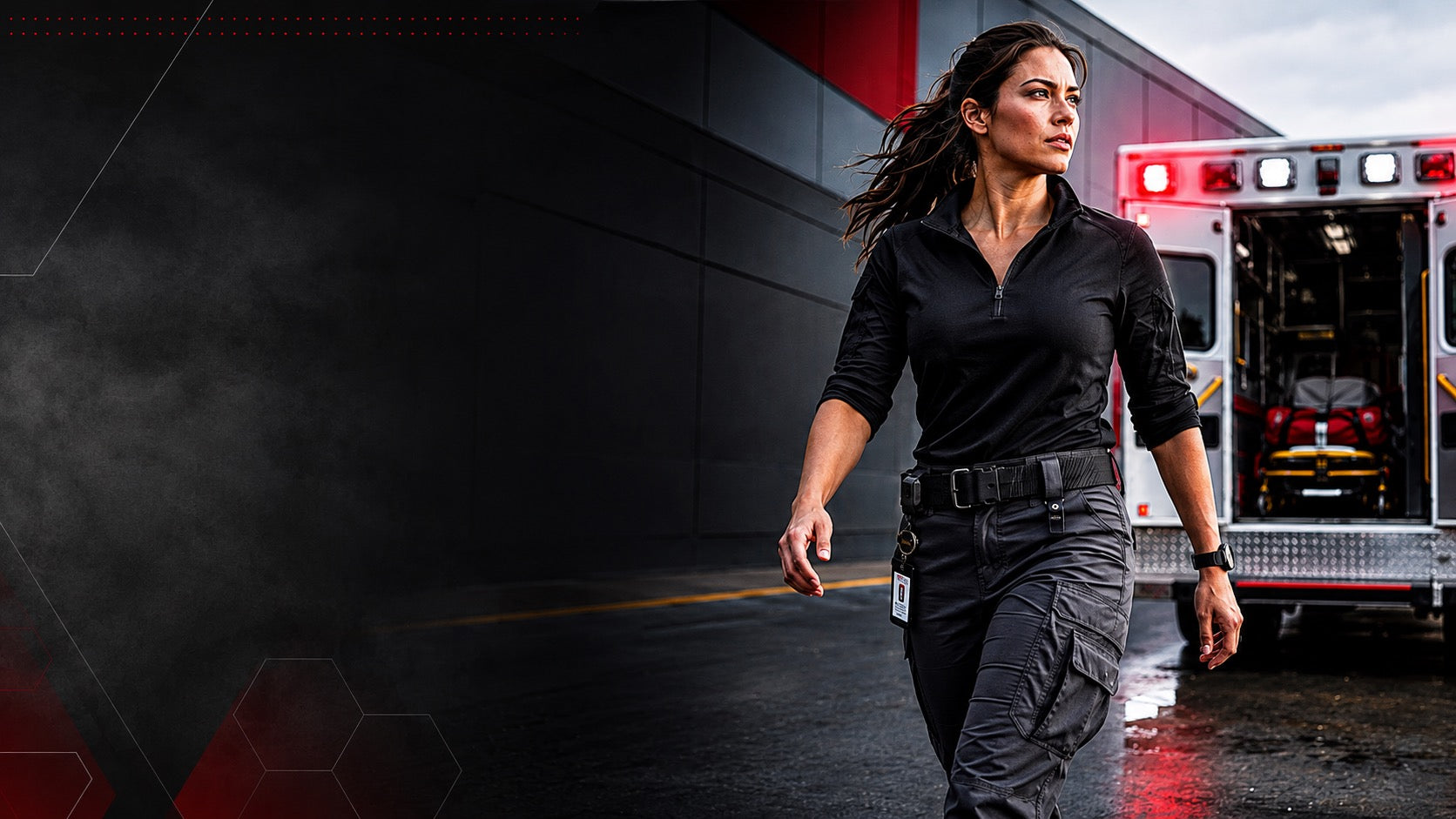 A woman in a black tactical-style shirt and dark cargo pants walks in front of an ambulance with flashing lights, with a dark industrial building in the background.