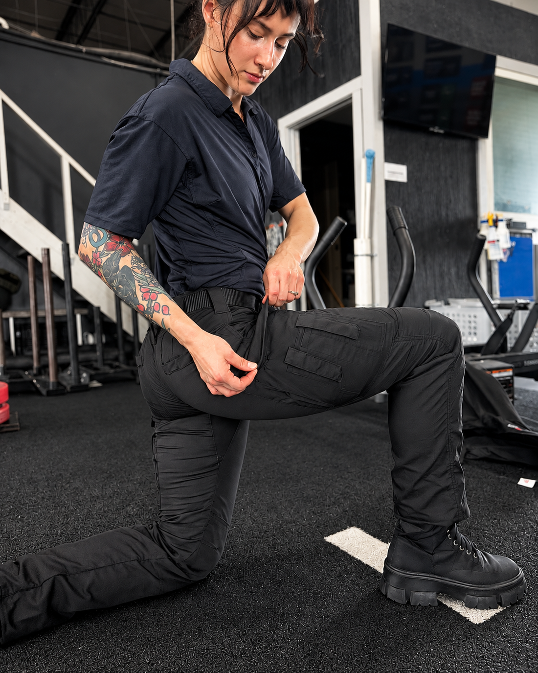 Woman kneeling in a gym wearing black MGT Gear tactical pants and demonstrating the cargo pocket